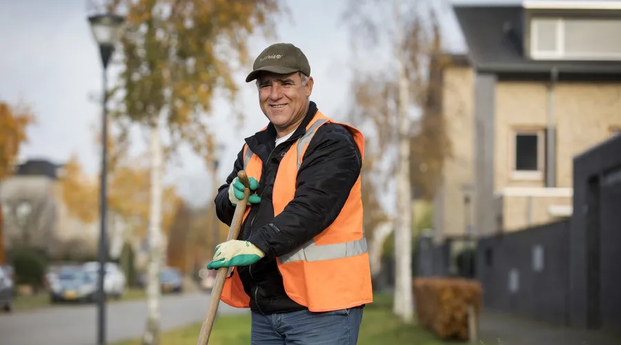 Vergunninghouder werkt in het groen en heeft een schoffel in zijn handen en kijkt lachend de camera in