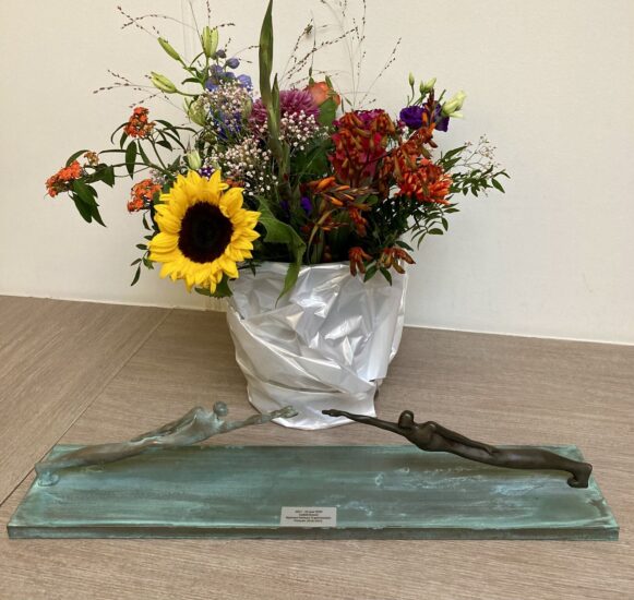 Bloemen, en daarvoor het kunstwerk ‘De nieuwe toekomst’ met twee figuren die met de hand naar elkaar toe reiken, om elkaar te vinden.